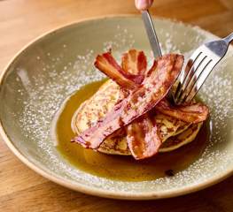 A knife and fork cutting into a stack on pancakes topped with streaky bacon and maple syrup