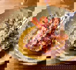 A knife and fork cutting into a stack on pancakes topped with streaky bacon and maple syrup