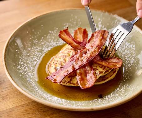A knife and fork cutting into a stack on pancakes topped with streaky bacon and maple syrup