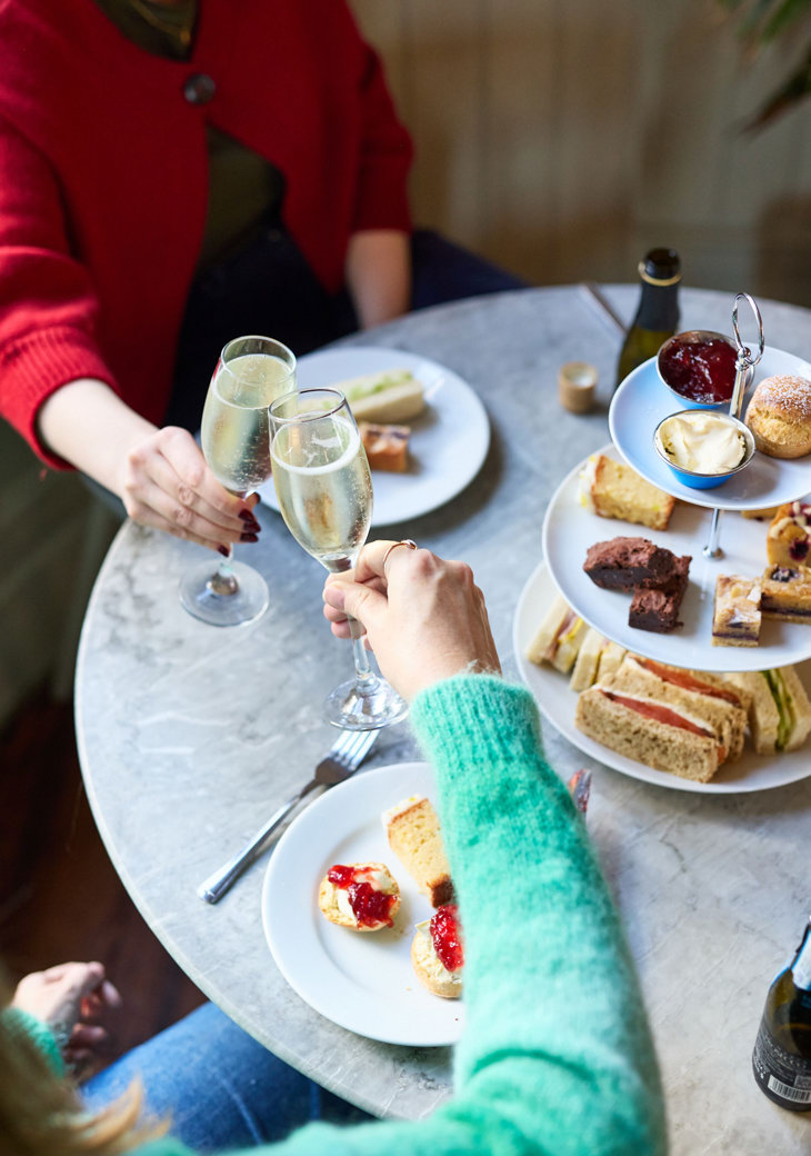 Two hands clinking glasses of prosecco over a table laden with an afternoon tea spread of finger sandwiches, mini cakes, scones and tea.