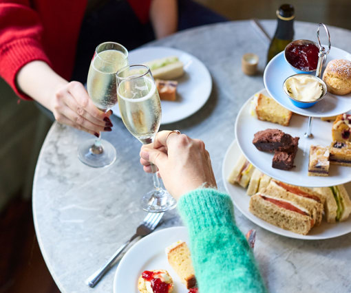 Two hands clinking glasses of prosecco over a table laden with an afternoon tea spread of finger sandwiches, mini cakes, scones and tea.