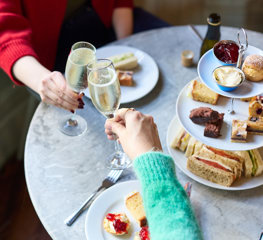 Two hands clinking glasses of prosecco over a table laden with an afternoon tea spread of finger sandwiches, mini cakes, scones and tea.