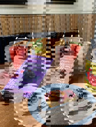 A hand holding a book with a delicious slice of cake and a selection of ice cold drinks on the table