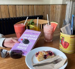 A hand holding a book with a delicious slice of cake and a selection of ice cold drinks on the table