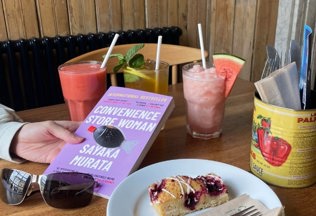 A hand holding a book with a delicious slice of cake and a selection of ice cold drinks on the table