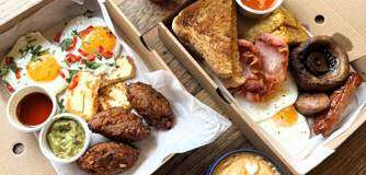 Two takeaway boxes filled with dishes. Our Sweetcorn Hash on the left and class Boss Breakfast on the right. A coffee and a smoothie in takeaway containers also sit on the table.