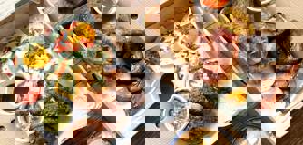 Two takeaway boxes filled with dishes. Our Sweetcorn Hash on the left and class Boss Breakfast on the right. A coffee and a smoothie in takeaway containers also sit on the table.