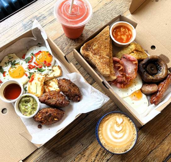Two takeaway boxes filled with dishes. Our Sweetcorn Hash on the left and class Boss Breakfast on the right. A coffee and a smoothie in takeaway containers also sit on the table.