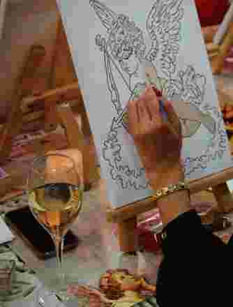 A woman's had paints an angel onto a canvas with a glass of wine nearby