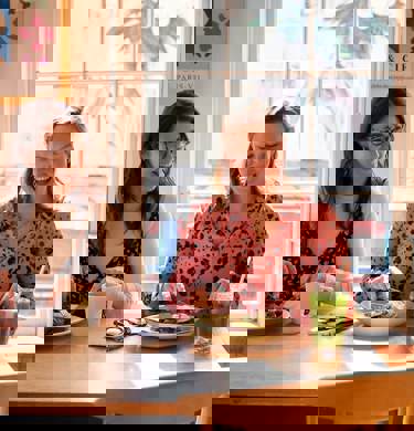 Two women, sitting next to one another, smiling and enjoying brunch dishes and drinks together