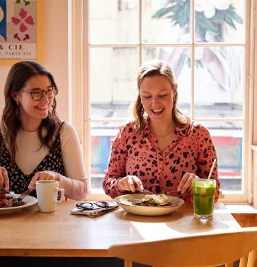 Two women, sitting next to one another, smiling and enjoying brunch dishes and drinks together