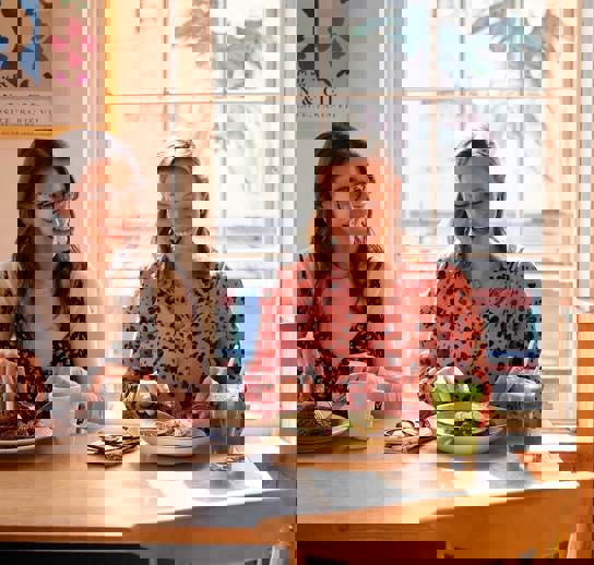 Two women, sitting next to one another, smiling and enjoying brunch dishes and drinks together