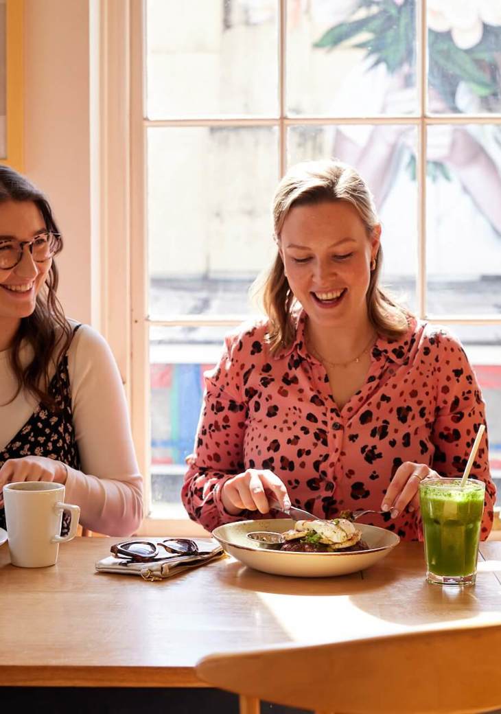 Two women, sitting next to one another, smiling and enjoying brunch dishes and drinks together