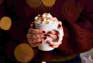 two hands with burgundy painted nails clasp around a hot chocolate filled with whipped cream and marshmallows. The person holding the cup wears a cosy burgundy jumper.