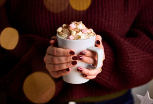 two hands with burgundy painted nails clasp around a hot chocolate filled with whipped cream and marshmallows. The person holding the cup wears a cosy burgundy jumper.