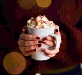 two hands with burgundy painted nails clasp around a hot chocolate filled with whipped cream and marshmallows. The person holding the cup wears a cosy burgundy jumper.