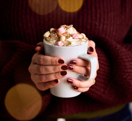 two hands with burgundy painted nails clasp around a hot chocolate filled with whipped cream and marshmallows. The person holding the cup wears a cosy burgundy jumper.