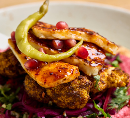 A close up shot of a flatbread with colourful houmous, slaw, cauliflower and halloumi on top with a thin, green chilli pepper