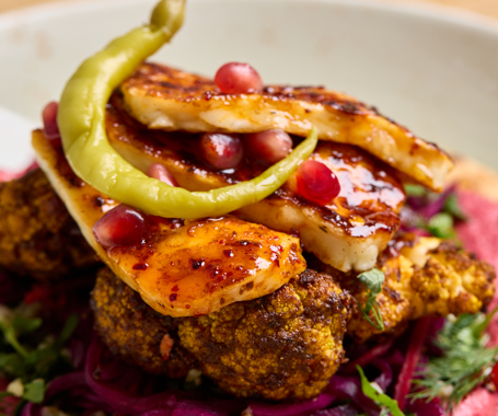 A close up shot of a flatbread with colourful houmous, slaw, cauliflower and halloumi on top with a thin, green chilli pepper