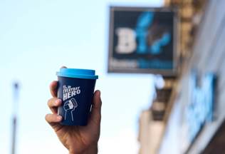 A hand holding a blue reusable coffee cup with the words 'Be an everyday hero' in white written across it